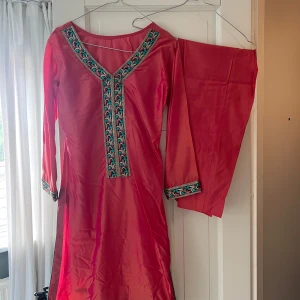Rosa klänning från Pakistan side klänning med broderi med byxor till - Säljer en pakistansk klänning med v-ringning och vackra broderade detaljer i blått och svart längs halsringningen och ärmsluten. Klänningen har långa ärmar och är tillverkad i ett glansigt silke material som ger en lyxig känsla. Kommer med tillhörande byxor.