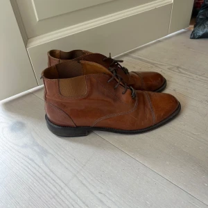 Bruna skinnkängor med snörning - Snygga eleganta bruna kängor i skinn från Alfred Sargent med klassisk snörning och låg klack. Skorna har rund tå och elastisk panel vid ankeln för extra komfort. Perfekta för dig som gillar en stilren och tidlös look.