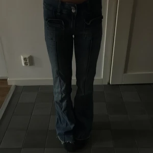 Blå bootcut jeans med fickdetaljer - Säljer ett par blå jeans med bootcut-modell och snygga fickor framtill. Jeansen har hög midja och råa kanter nertill för en cool look. Perfekta för dig som gillar en avslappnad men trendig stil.