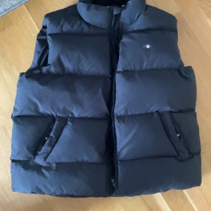 Gant Dunväst - Säljer en svart, tjock dunväst med hög krage och två snedställda fickor framtill. Västen har en clean look med breda, quiltade paneler och stängs med dragkedja. Perfekt att slänga över en hoodie när det är kyligt ute.