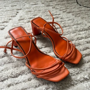 Orange sandaler med klack och remmar - Snygga orange sandaler från Star Collection med fyrkantig tå, smala remmar och blockklack. Skorna har en trendig design med knytning runt vristen och är tillverkade i syntetmaterial. Perfekta för att lyfta din sommarlook.