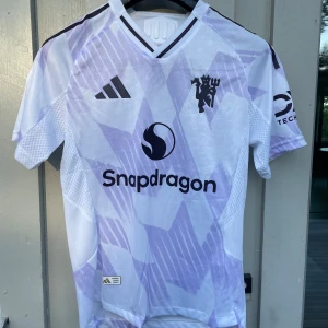 Manchester United bortatröja Adidas S - Säljer en Manchester United bortatröja från Adidas i storlek S. Tröjan är vit med ljuslila geometriskt mönster, svart klubbmärke och sponsorlogga. Tillverkad i ventilerande polyester med korta ärmar och slim fit passform. Perfekt för dig som älskar fotboll och vill sticka ut.
