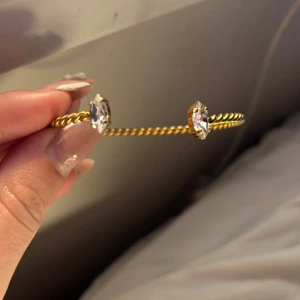 Guldigt armband med kristaller - Snyggt armband i guldton med tvinnad design och två stora, gnistrande kristaller i varje ände. Perfekt accessoar för att lyfta din outfit med lite extra bling och stil.