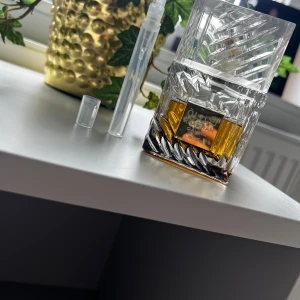 Lattafa Khamrah Qahwa parfym - Lattafa Khamrah Qahwa parfym i en lyxig, transparent glasflaska med mönstrad design och fyrkantigt lock. Vätskan är bärnstensfärgad och flaskan har en guldfärgad etikett. Medföljer en liten genomskinlig sprayflaska för decant.