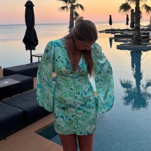 populär adoore klänning - Superfin kort klänning i grönt med blommigt mönster och v-ringning. Klänningen har långa puffärmar och knappar framtill, perfekt för sommarkvällar. Sitter snyggt i midjan och har en fräsch vibe.