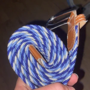 Blåvitt flätat bälte  - Snyggt flätat bälte i blått och vitt med bruna skinn-detaljer vid spännet. Bältet har ett klassiskt silverfärgat spänne och är gjort i ett mjukt, flexibelt material. Perfekt för att ge din outfit en fräsch och sportig touch väldigt nytt använd några gånger