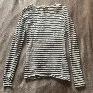 Randig långärmad tröja - Säljer en klassisk långärmad tröja med smala marinblå och vita ränder. Tröjan har en rund halsringning och är figurnära i modellen. Perfekt att styla med jeans eller kjol för en enkel och stilren look.