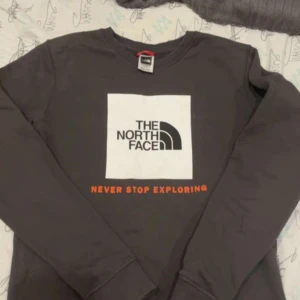 Svart långärmad tröja från The North Face - Säljer en svart långärmad tröja från The North Face med vit logga och texten 'Never Stop Exploring' i orange framtill. Tröjan har rund halsringning och är perfekt för en avslappnad stil.