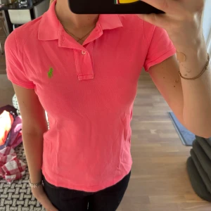 Rosa pikétröja från Ralph Lauren - Säljer en klassisk rosa pikétröja från Ralph Lauren med grönt broderat logotyp på bröstet. Tröjan har krage, korta ärmar och knappar framtill. 