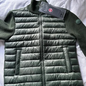 Grön Cardigan från Colmar - Snygg grön pufferjacka från Colmar med stickade ärmar och krage. Jackan har dragkedja framtill och två fickor med dragkedja. Perfekt för kyliga dagar och ger en modern look med sin kombination av material.