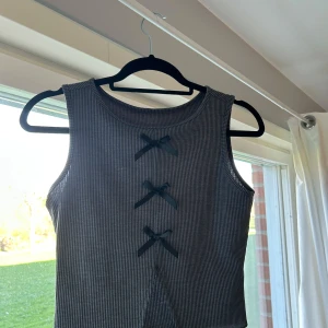 Ribbad grå croppad linnetopp med rosetter - Säljer en grå ribbad croppad linnetopp med tre svarta rosetter framtill. Toppen är ärmlös och har en rund halsringning. Har även en cutout nedtill.