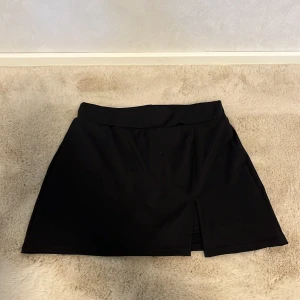 Svart kortkjol med slits - Säljer en svart minikjol med en liten slits framtill. Kjolen har en bred linning och är sydd i ett stretchigt material. Under kjolen finns inbyggda shorts för extra komfort och rörelsefrihet. (Blå minikjol på köpet)