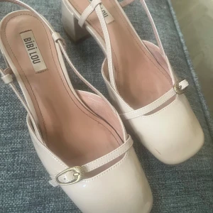 Beige klackskor från BiBi Lou - Säljer ett par stilrena beige klackskor från BiBi Lou med fyrkantig tå och justerbara remmar med spänne. Skorna har en låg, bred klack och är tillverkade i syntetmaterial med glansig finish.