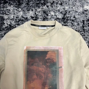 Beige sweatshirt från Calvin Klein med tryck - Säljer en beige sweatshirt från Calvin Klein med ett stort konstnärligt tryck i rött, svart och rosa på bröstet. Tröjan har rund halsringning och långa ärmar. Perfekt för en avslappnad och trendig stil.