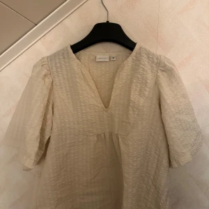 Beige blus med puffärmar från vila - Säljer en beige blus från vila med v-ringning och puffiga trekvartsärmar. Blusen har ett lätt strukturerat tyg och en lös passform, perfekt för en stilren look.