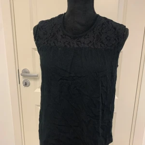 Svart ärmlös blus med spetsdetalj - Svart ärmlös blus med vacker spets upptill över axlar och bröst. Blusen har rund halsringning och är tillverkad i ett lätt, mjukt tyg. Perfekt för en stilren och elegant look.