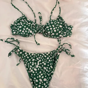 Grön bikini med vita prickar - Snygg grön bikini med vita prickar och knytband både upptill och nedtill. Överdelen har vadderade kupor och bygel, och underdelen har höga ben och knyt i sidorna. Perfekt för stranden eller poolen!