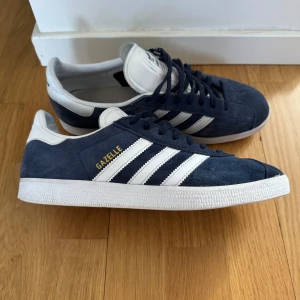 Adidas Gazelle blå sneakers - Säljer ett par klassiska Adidas Gazelle sneakers i mörkblå mocka med vita detaljer och de ikoniska tre ränderna på sidorna. Skorna har vit sula och snörning framtill. Perfekta för en stilren och sportig look.