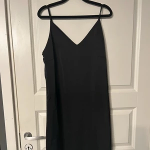 Svart långklänning med spetskant - Säljer en svart långklänning med smala axelband och v-ringning. Klänningen har en elegant spetskant längst ner och är tillverkad i ett slätt, glansigt material som ger en lyxig känsla.