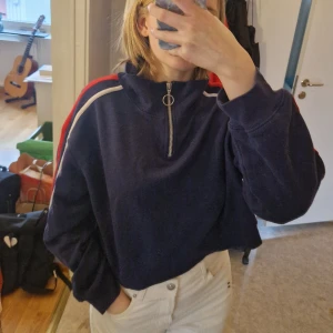 Blå croppad sweatshirt med dragkedja från H&M - Säljer en blå croppad sweatshirt från H&M Divided med dragkedja i storlek S. Tröjan har röda och vita ränder längs ärmarna.