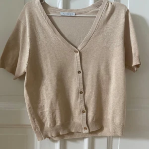 Beige kortärmad kofta från In Front - Säljer en beige kortärmad kofta från In Front med v-ringning och knappar framtill. Tröjan är i mjukt material och har ribbade kanter vid ärmslut och nederkant. Perfekt att slänga på sig över ett linne ex. Jag köpte den i second hand butik och har tvättat den och den är i så fint skick men inte använt särskilt mycket och testar därför att sälja den 😇😇😛😛