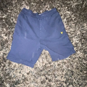 Mörkblå shorts från Lyle & Scott - Mörkblå shorts med resår i midjan och en gul broderad logga från Lyle & Scott på vänster ben. Shortsen har en bakficka och är tillverkade i mjukt bomullsmaterial. Perfekta för en avslappnad stil. Obs storlek stämmer inte det är för 14-15 år gamla