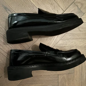 Svarta loafers från Pavement - Säljer ett par klassiska svarta loafers i modellen Naeli från Pavement. Skorna har en stilren design med rund tå och platt sula, tillverkade i blankt skinn. Perfekta för dig som gillar en tidlös och clean look.