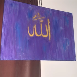 Lila tavla med gul arabisk text - Säljer en handmålad tavla med lila bakgrund och gul text på arabiska som betyder 'Allah'. Tavlan är målad på duk och har en abstrakt känsla med inslag av blått. Perfekt som väggdekoration.