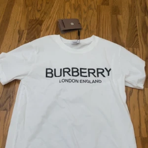 Vit t-shirt från Burberry - Vit t-shirt från Burberry med klassisk svart logotyp 'BURBERRY LONDON ENGLAND' tryckt framtill. T-shirten har rund halsringning och korta ärmar. Perfekt för en stilren och trendig look.