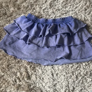 Blå volangkjol från Monki - Supersöt blålila kjol från Monki med dubbla volanger och resår i midjan. Kjolen är kort och har en mjuk känsla tack vare materialblandningen av bomull och polyester. Perfekt för dig som gillar en lekfull och trendig stil.