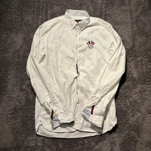 Morris - Klassisk vit skjorta från Morris med broderad logga på bröstet och knappar framtill. Skjortan har lång ärm och diskreta randiga detaljer på insidan av kragen och manschetterna. Perfekt för en stilren look.