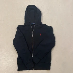  Ralph Lauren Hoodie - Säljer en svart hoodie från Ralph Lauren med dragkedja och en liten röd logotyp på bröstet. Perfekt för en avslappnad look. Dragkedjan är dock lätt att åka ner. Och en liten slitning längst ut på ärmen 