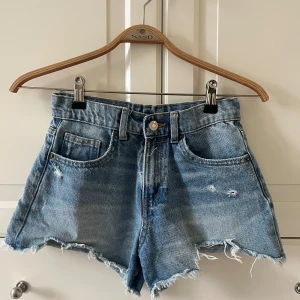 Blå jeansshorts med fransar från Zara - Säljer ett par klassiska blå jeansshorts från Zara med fransig kant och slitna detaljer.Perfekta för sommaren och enkel att matcha med olika toppar och i Bra skick!