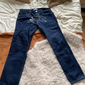 Mörkblå jeans med kattmotiv från BBC Ice Cream - Unika mörkblå jeans från BBC Ice Cream med ett coolt kattmotiv i nitar framtill. Klassisk femficksmodell med raka ben och kontrastsömmar. Perfekta för dig som vill sticka ut med din stil.