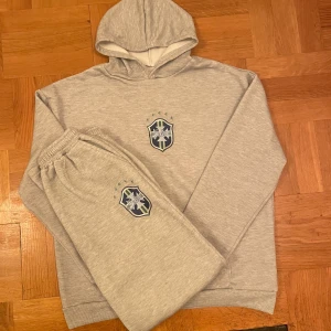 Grå tracksuit hoodie och byxor - Säljer en ljusgrå  tracksuit alltså hoodie och byxorna, båda med Brasil-inspirerat tryck på bröstet det är klistrat på och sen på benet. Den är perfekt för en avslappnad stil, den är jätte bekväm och jätte stilig. Den är helt ny har aldrig andvänt den bara testad. Den är storlek L fast den kan passa ganska stor. Priset går att diskuteras vid ett snabb äffär. Vid frågor är det bara fråga:)