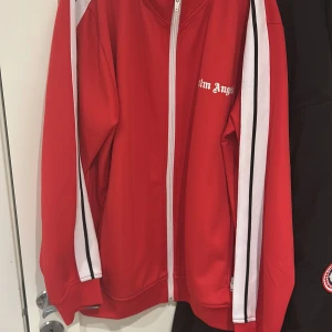Röd track jacket från Palm Angels - Röd track jacket från Palm Angels med vita och svarta ränder längs ärmarna och vit dragkedja framtill. Jackan har hög krage och Palm Angels-logga tryckt på bröstet. Perfekt för en sportig och trendig look. Anväd max 10 gånger fins tag 2000kr kan sänka vid snabb affär