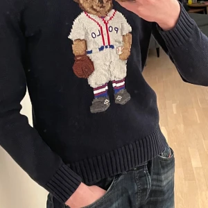 Mörkblå stickad tröja med björn från Polo Ralph Lauren - Säljer en mörkblå stickad tröja från Polo Ralph Lauren med deras ikoniska björn i basebollkläder på framsidan. Tröjan har rund halsringning, långa ärmar och ribbade muddar. Dessvärre finns ett mindre hål på vänster underarm (se bild 5). Tröjan är egentligen barn storlek 16 år men passar lika bra som XS vuxen storlek 