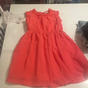 Korallrosa kortklänning med volangdetaljer - Säljer en söt korallrosa klänning med volang vid ärmslut och halsringning. Klänningen har en markerad midja och är tillverkad i ett lätt, luftigt material. Perfekt för varma dagar och somriga tillfällen.