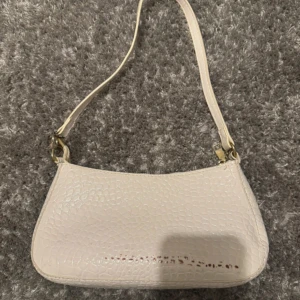 Ljusbeige/vit axelväska med krokodil mönster - Säljer en stilren ljusbeige axelväska med krokopräglat mönster och guldfärgade detaljer. Väskan har dragkedja upptill och ett rymligt svart innerfoder. Perfekt för att bära det viktigaste till vardags.🤍