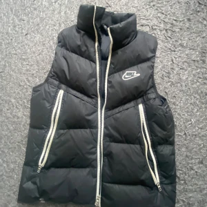 Svart dunväst från Nike - Snygg svart dunväst från Nike med hög krage och två dragkedjefickor framtill. Västen har en vit Nike-logga på bröstet och tydliga quiltade partier. Perfekt för lager-på-lager-stil under kyliga dagar. Riktigt bra skick och rare. Nypris 1905kr
