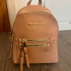 Ljusrosa ryggsäck från Michael Kors - Säljer en ljusrosa ryggsäck från Michael Kors med guldfärgade detaljer och dragkedjor. Väskan har ett huvudfack och en mindre ficka framtill, båda med dragkedja. Perfekt storlek för vardagens alla prylar.