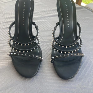 Svarta klackar med nitar från Zara  - Säljer ett par svarta klackar från Zara Trafaluc med flera remmar dekorerade med silvriga nitar. Skorna har öppen tå och hög klack, perfekta för att ge outfiten edge. Snygg och trendig design!