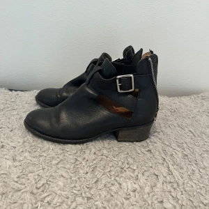 Svarta sandaler / boots med spänne och dragkedja - Säljer ett par svarta boots / sandaler med coola detaljer som spänne på sidan och dragkedja baktill. Skorna har låg klack och rund tå, samt öppna partier på sidorna.