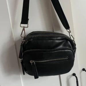 Svart axelväska med dragkedjor - Säljer en svart axelväska från Carin Wester i skinnimitation med flera fack och silverfärgade dragkedjor. Väskan har en bred, justerbar axelrem och minimalistisk design. Perfekt för att bära det viktigaste med stil. Har använts en del så har lite slitningar i sömmarna men håller fortfarande väldigt bra. Endast att det ser slutet ut när man kollar nära, inte på längre håll. Skriv för fler bilder eller frågor💞