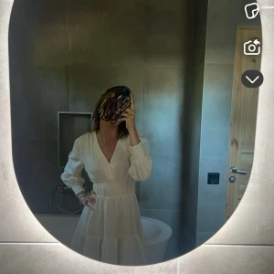 Vit studentklänning med volang - Superfin vit klänning med v-ringning och långa ärmar. Smockad midja som ger en snygg siluett och volangdetaljer nertill. Perfekt för sommarens alla tillfällen eller studenten. 