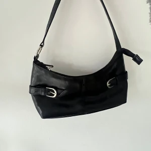 Svart axelväska med spänndetaljer - Snygg svart axelväska i skinnimitation med två dekorativa spännen framtill. Väskan har en enkel, stilren design och ett justerbart axelband. Perfekt för att bära dina viktigaste saker till vardags eller när du vill ha en klassisk look.💕