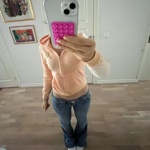 Persikofärgad hoodie från Juicy Couture - Säljer en snygg persikofärgad hoodie från Juicy Couture med dragkedja och logga på bröstet. Tröjan har långa ärmar och en mysig huva. Perfekt för en avslappnad stil.