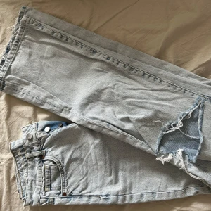 Ljusblå wide leg jeans med slitningar - Säljer ett par ljusblå jeans med breda ben och slitna detaljer på båda benen. Byxorna har knappar i gylfen och klassisk femficksdesign. Modellen är regular wide med midja 26 och längd 32.