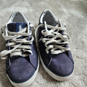 Blå sneakers från Polo Ralph Lauren - Säljer ett par blå sneakers från Polo Ralph Lauren med vit logga på plösen och vit sula. Skorna har vita snören och klassisk låg profil. Perfekta för en avslappnad stil.