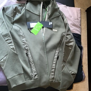 Grön jacka från BOSS med dragkedja och byxa  - Snygg grön jacka och byxa från BOSS med dragkedja framtill och längs fickorna. Jackan har en sportig look med diskreta detaljer och logga på bröstet. Perfekt för dig som gillar stilrena plagg med modern känsla.